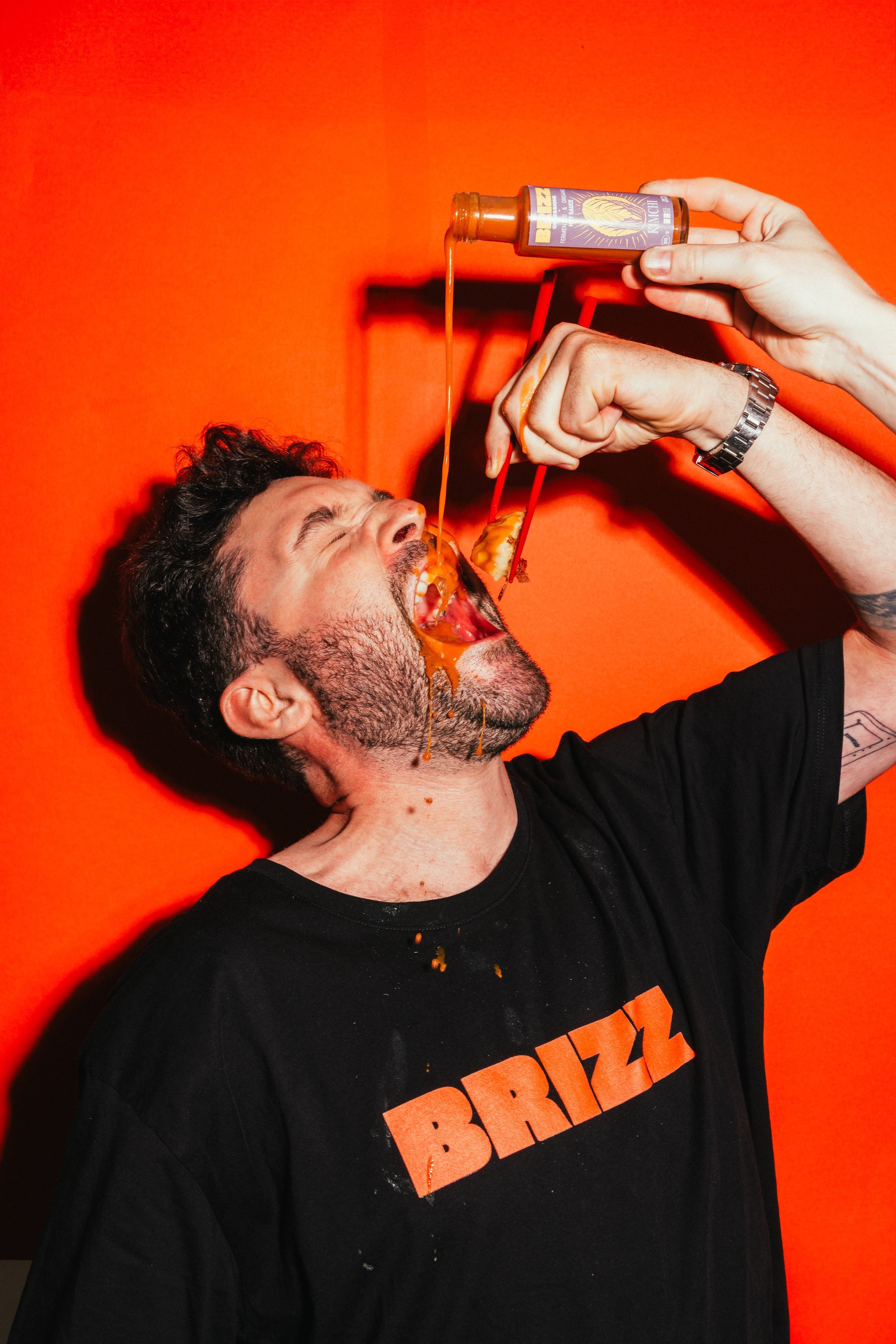 Johannes Schartner genießt BRIZZ Kimchi Hot Sauce direkt aus der Flasche vor oranger Wand.