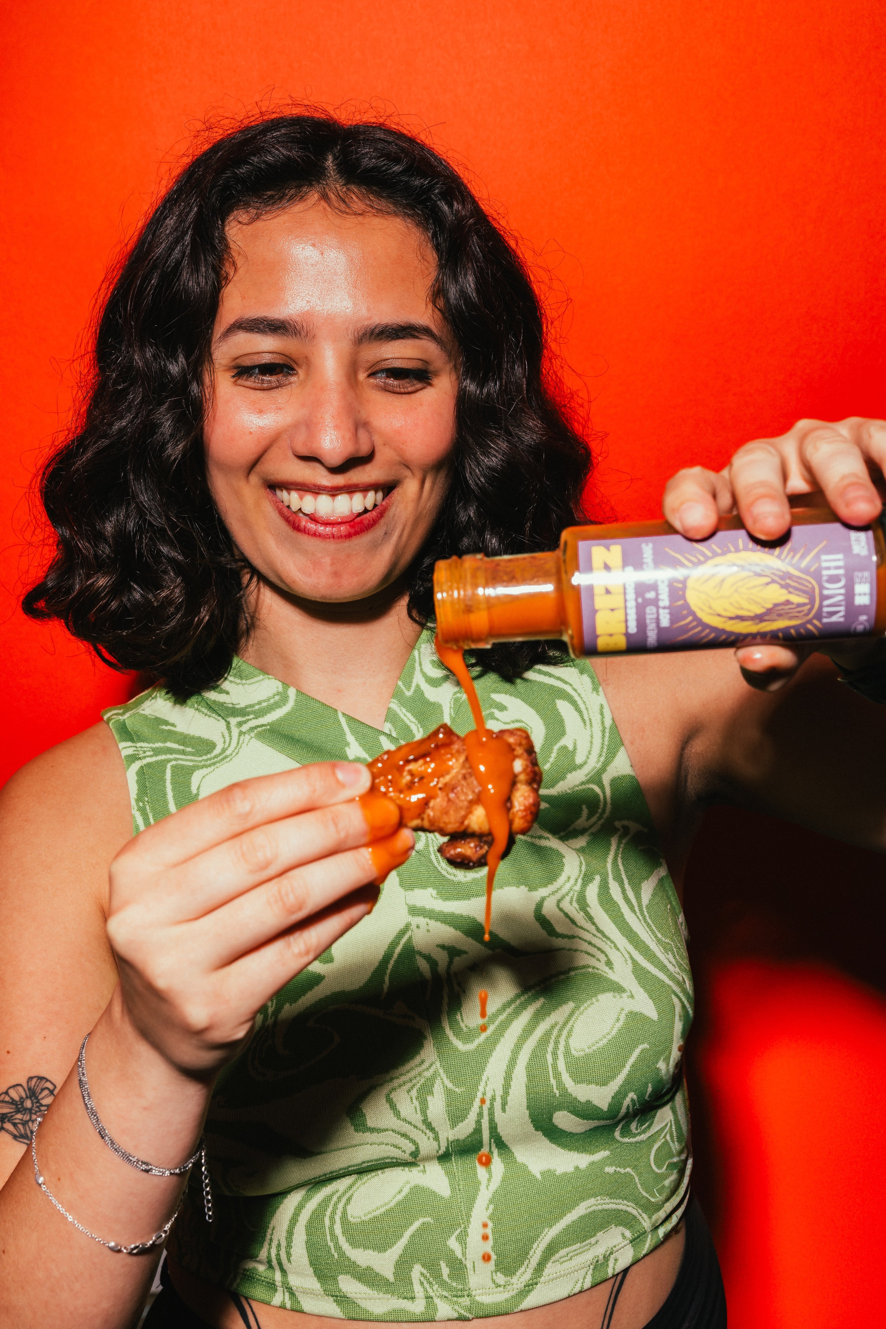 Frau lacht und träufelt BRIZZ Kimchi Hot Sauce auf Chicken Wing – Bio, fermentiert, würzig.