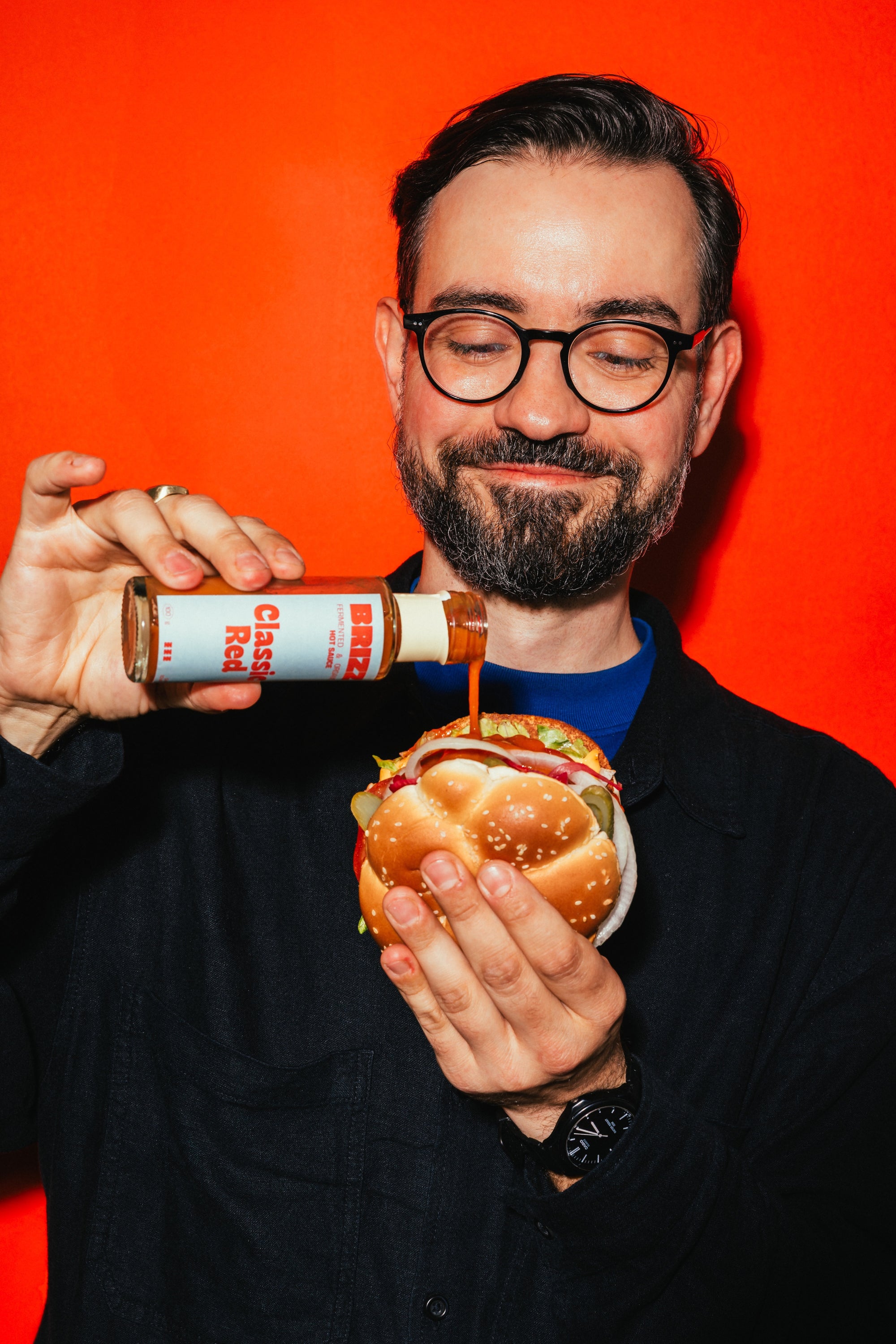 Ein Mann gießt BRIZZ Classic Red Hot Sauce über einen Burger vor oranger Wand.