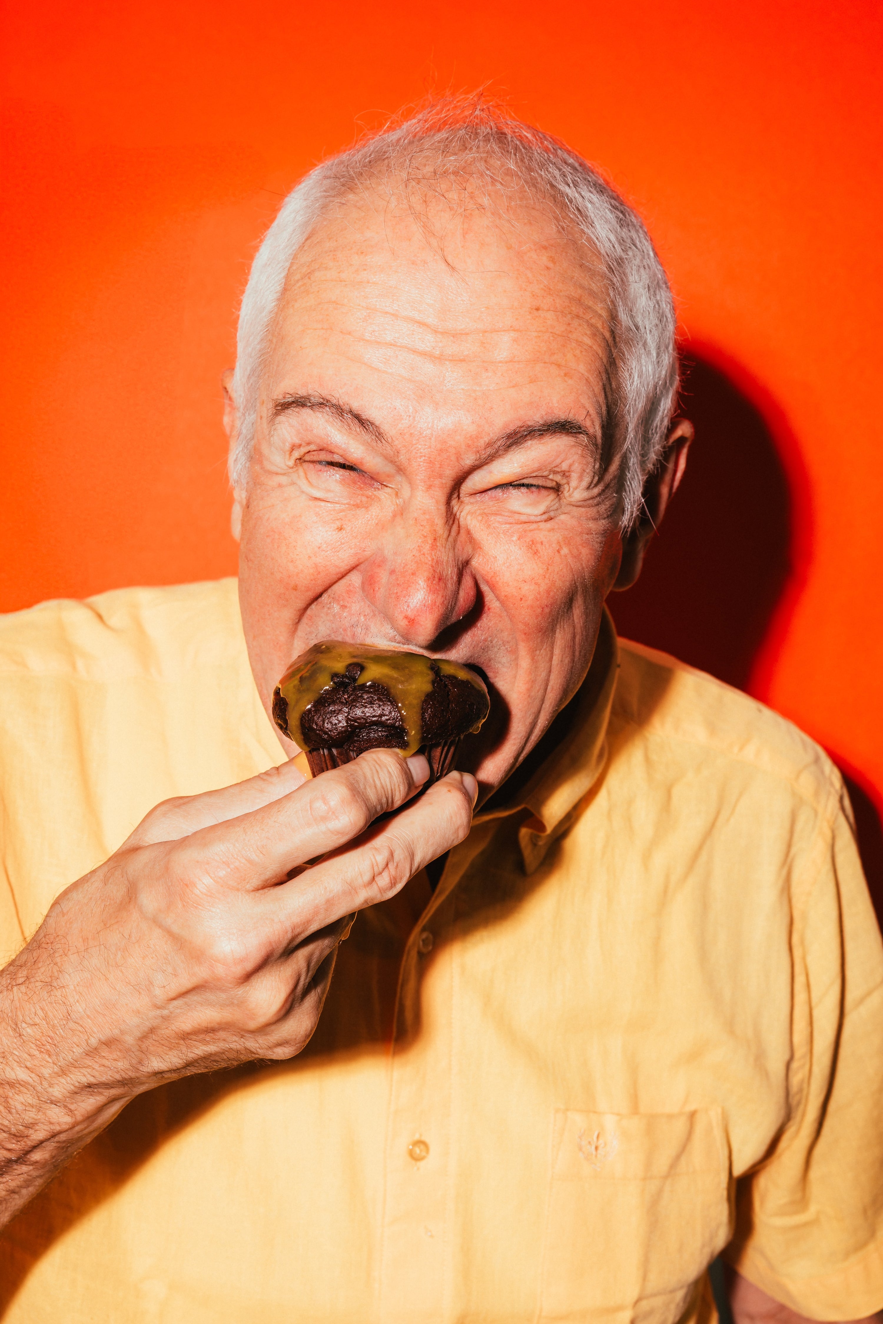 Ein älterer Mann genießt Schoko-Muffin mit BRIZZ Classic Red Hot Sauce vor leuchtend orangem Hintergrund.